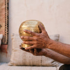 The maker's hands holding the perforated brass sphere of the Aman lamp