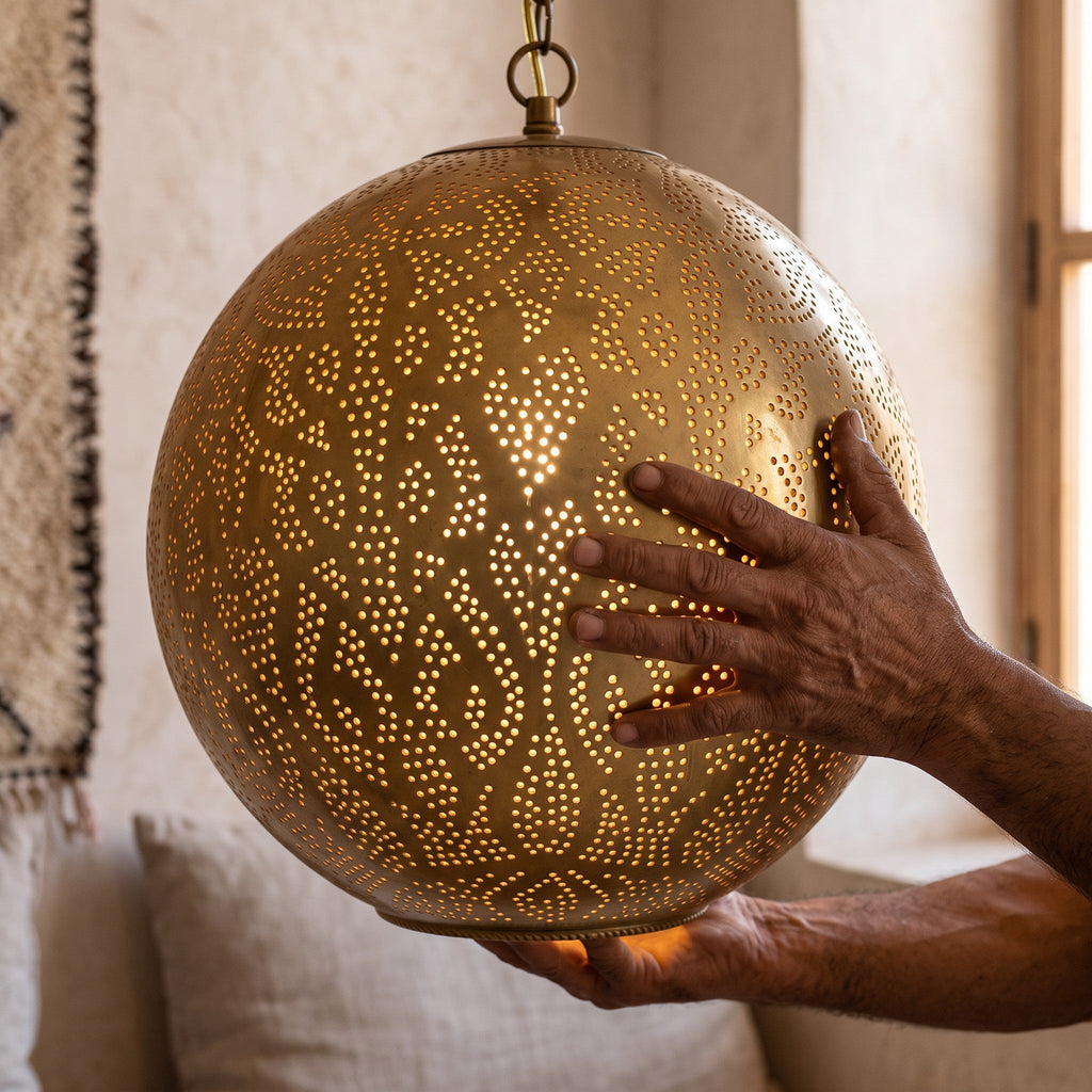 A maker's hands holding the pierced brass sphere of the Badr pendant in the workshop