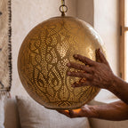 A maker's hands holding the pierced brass sphere of the Badr pendant in the workshop