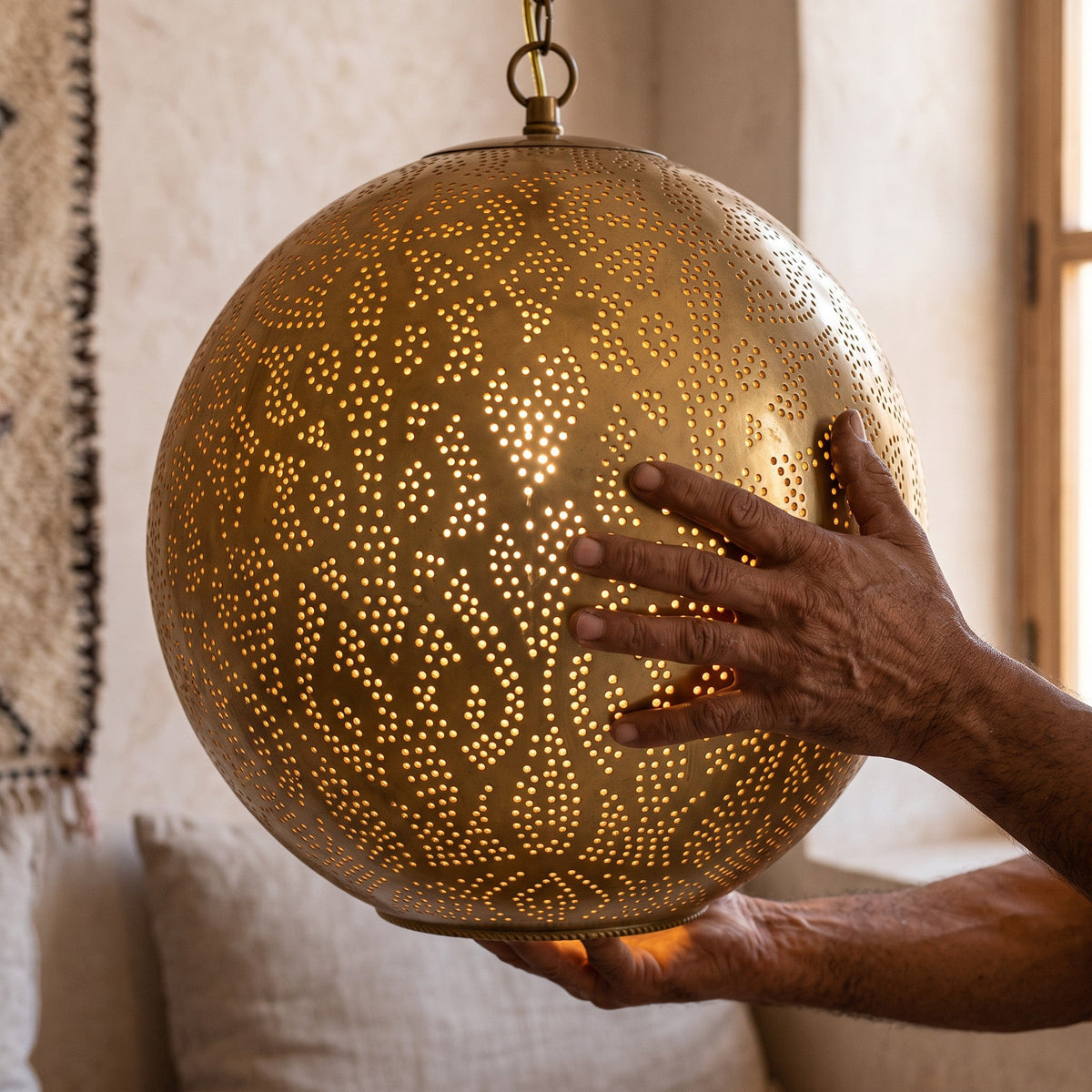 A maker's hands holding the pierced brass sphere of the Badr pendant in the workshop